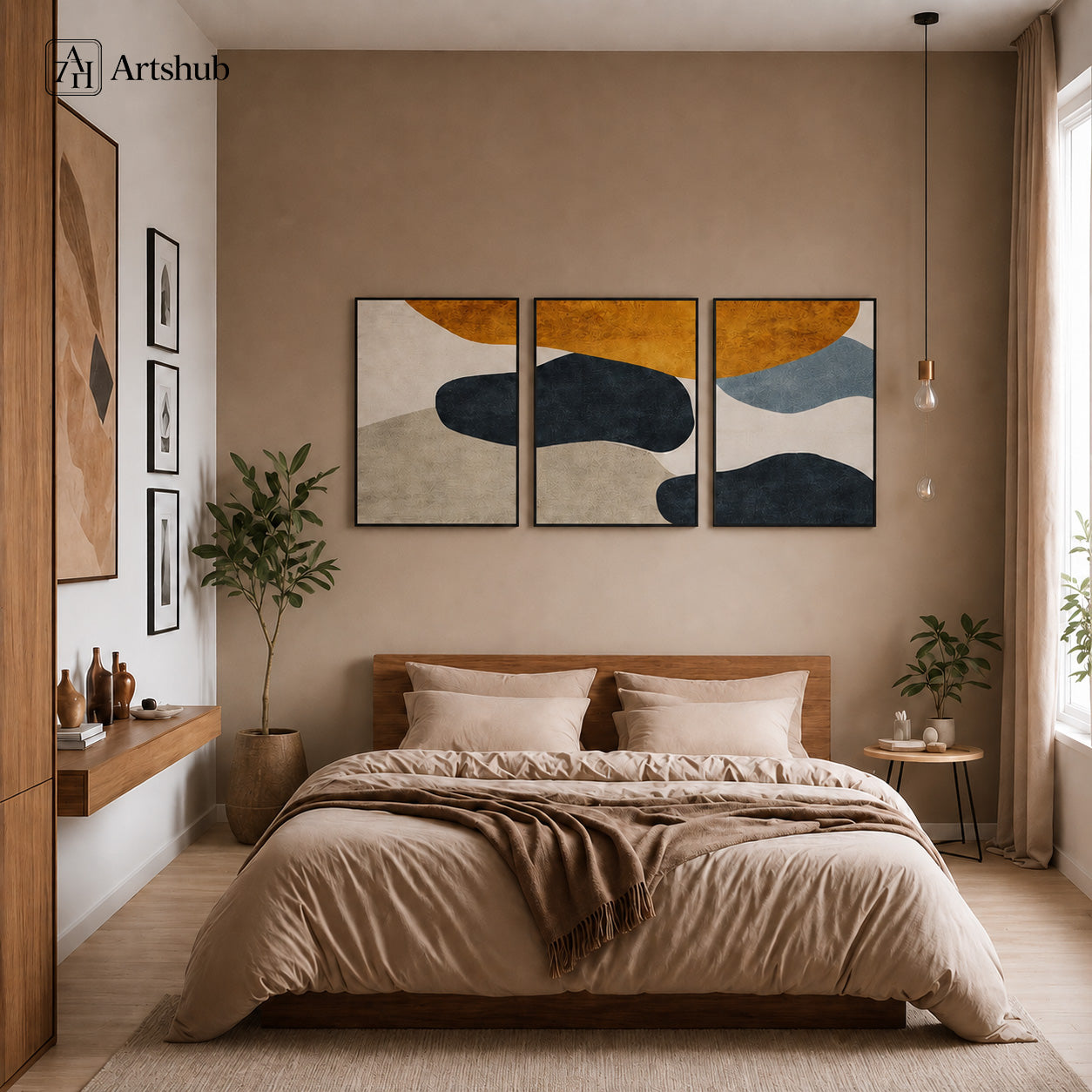 Artshub Amber Stone 3-panel triptych abstract canvas framed wall art displayed on a warm taupe bedroom wall above a wooden bed with cream linen bedding and brown throw, large green plant in terracotta pot, pendant bulb light, wooden shelving with ceramic vases and books, warm wood floor – bold abstract amber, navy and beige organic shapes in black floater frames