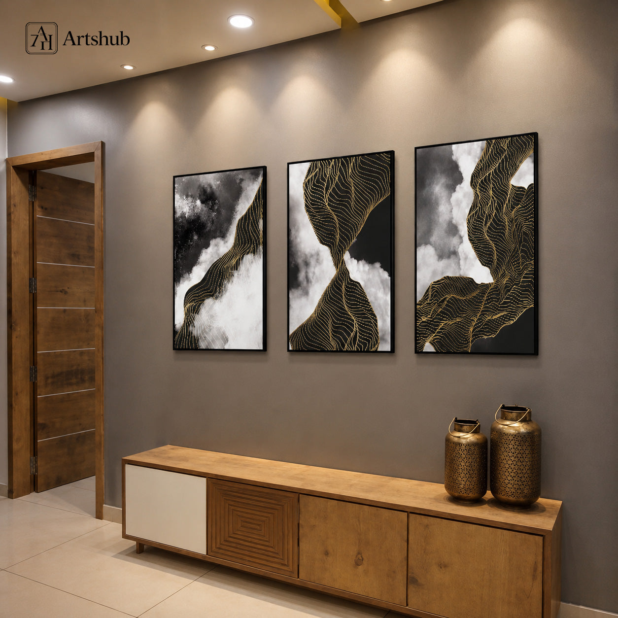 Artshub Obsidian Flow 3-panel triptych abstract canvas framed wall art displayed on a warm grey hallway wall above a wooden console sideboard with two gold metallic vases, recessed ceiling spotlights and wooden door – bold abstract black white and gold contour line triptych in premium black floater frames
